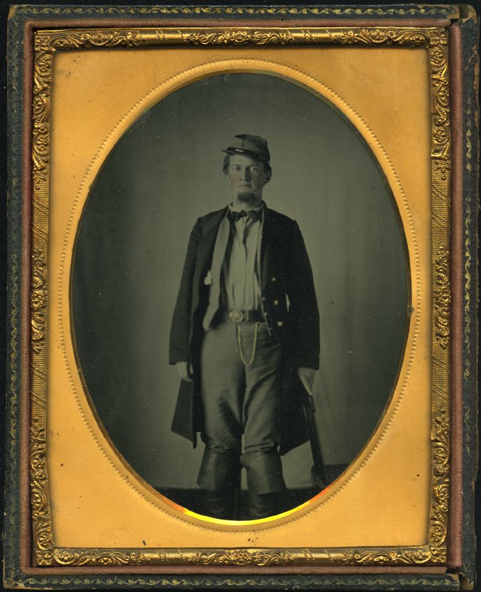 Half-plate ambrotype with applied color of a man, standing facing camera, looking at camera, in double-breasted uniform frock coat with three gold tinted buttons visible on subject's right side, waistocat open with three buttons visible on the right, and hankerchief handibng out of pocket on left side; shirt,with necktie and turned down collar, loose trousers with belt and round belt plate, watch chain at waist, cavalry boots, kepi, cocked to subject's right, short hair, goatee, cheeks and lips tinted pink; left arm hanging by side, right arm by side with right hand placed on the top of a spindle chair back, which is positioned to the side, plain back drop visible; oval gilt mat, embossed beading around oval; preserver stamped with foliate scroll design; gilt scrollwork in interior leather edging, embossed-leather-covered wood case with gem and foliate motif, door section of case missing, brass latch hooks intact.