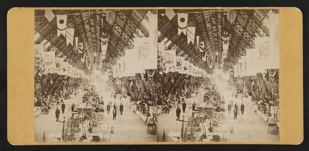 Historic stereoscope Photograph shows a large hall with flags hanging from the high ceiling, and people looking at the exhibits at the fair sponsored by the United States Sanitary Commission.
