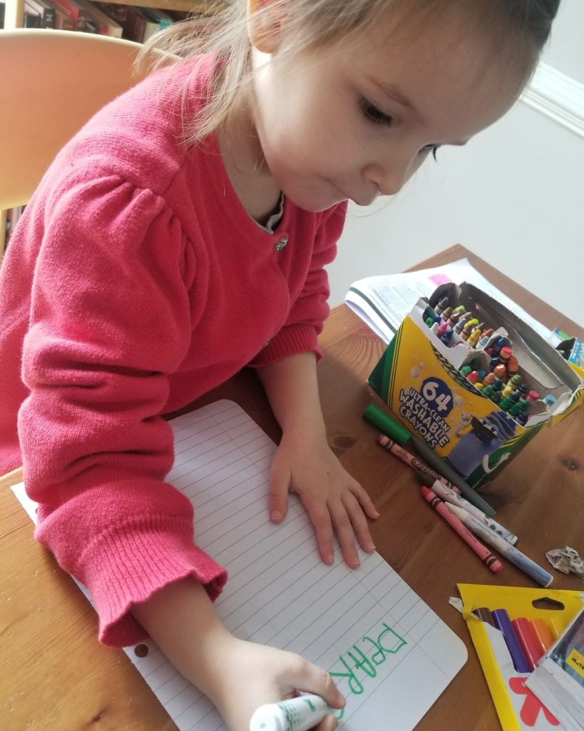 A toddler surrounded by crayons and markers writing a letter with a marker on lined paper.
