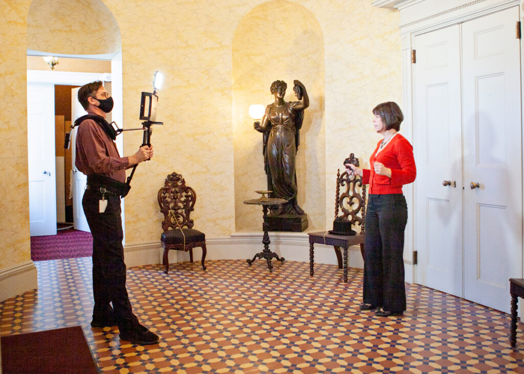 Two people are giving a virtual tour in the entrance hall of the White House of the Confederacy. 

There is a man on the left wearing glasses and a face mask holding a iPad on a shoulder rig made for filming, taping a woman on the right who has a brown bob haircut and wearing a red sweater. Behind the two people are two elaborate 19th Century chairs, a side table, and a statue of a Grecian woman holding a tragedy mask. 