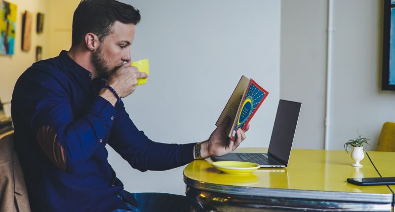 Man reading a book while drinking coffee at a computer table