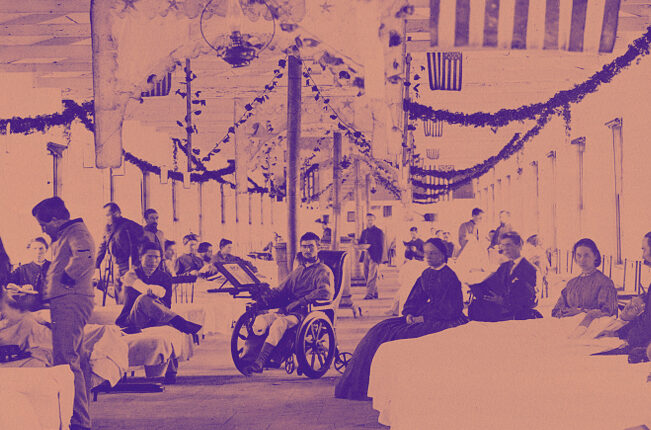 A Ward in Armory Square Hospital, Washington, D.C. U.S. soliders are lying in beds against the walls and one soldier is in a wheelchair in the center of the image. Two women nurses are seated next to bedridden soliders on the right. The ward's ceiling is decorated with streamers and U.S. Flags.