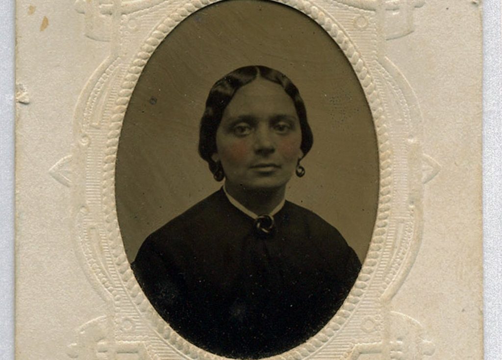 A tin type, black and white, portrait of Ellen Barnes. She is wearing a dark colored top with a white collar, wearing button-sized earrings, and her hair is up in a simple victorian style.